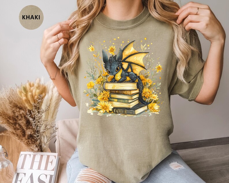Puede incluir: Camiseta caqui con una ilustraci&oacute;n de un drag&oacute;n negro con alas amarillas sobre una pila de libros, rodeado de flores amarillas. El dise&ntilde;o est&aacute; impreso en una camiseta c&oacute;moda e informal.