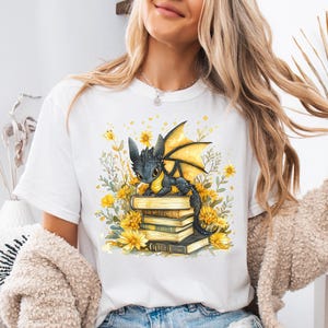Puede incluir: Camiseta blanca con una ilustraci&oacute;n en acuarela de un drag&oacute;n negro con alas amarillas posado sobre una pila de libros. El dise&ntilde;o est&aacute; rodeado de flores amarillas y vegetaci&oacute;n.