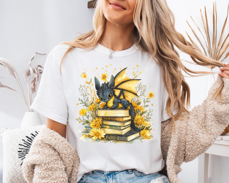 Puede incluir: Camiseta blanca con una ilustraci&oacute;n en acuarela de un drag&oacute;n negro con alas amarillas posado sobre una pila de libros. El dise&ntilde;o est&aacute; rodeado de flores amarillas y vegetaci&oacute;n.