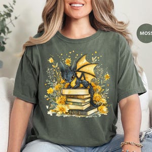 Puede incluir: Camiseta verde musgo con una ilustraci&oacute;n fant&aacute;stica. Muestra un drag&oacute;n negro con alas amarillas sobre una pila de libros, rodeado de flores amarillas y follaje. El dise&ntilde;o tiene un aire caprichoso y de cuento de hadas.