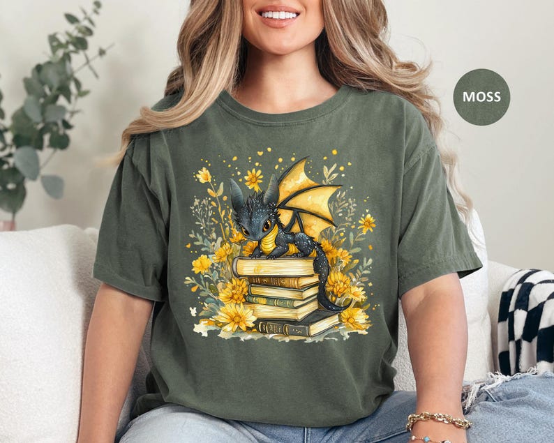 Puede incluir: Camiseta verde musgo con una ilustraci&oacute;n fant&aacute;stica. Muestra un drag&oacute;n negro con alas amarillas sobre una pila de libros, rodeado de flores amarillas y follaje. El dise&ntilde;o tiene un aire caprichoso y de cuento de hadas.