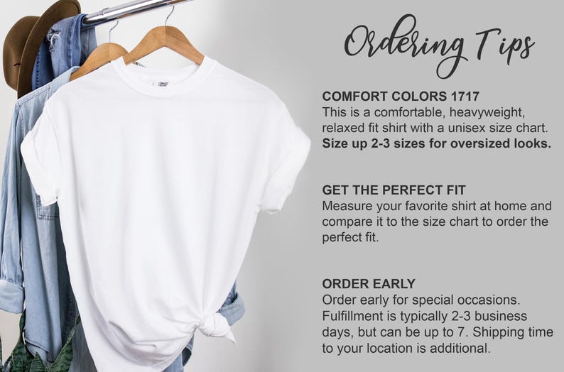 Puede incluir: Una camiseta blanca Comfort Colors 1717 cuelga de una percha de madera. La camiseta forma parte de una tabla de tallas unisex y tiene un corte relajado. La imagen tambi&eacute;n incluye una camisa de mezclilla y un sombrero marr&oacute;n.