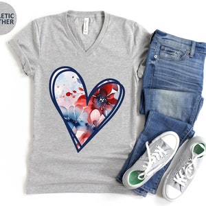 May include: A gray V-neck t-shirt with a blue outlined heart design. The heart is filled with a watercolor pattern of red, blue, and white flowers.