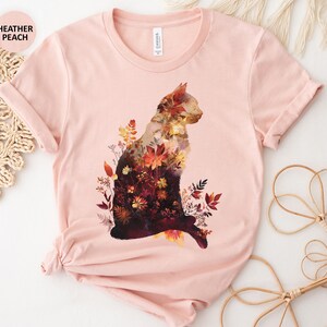May include: Heather peach t-shirt featuring a cat silhouette filled with autumn flowers and foliage in shades of red, orange, and yellow. The shirt has short sleeves and a crew neck. The text "HEATHER PEACH" is in the upper left corner.