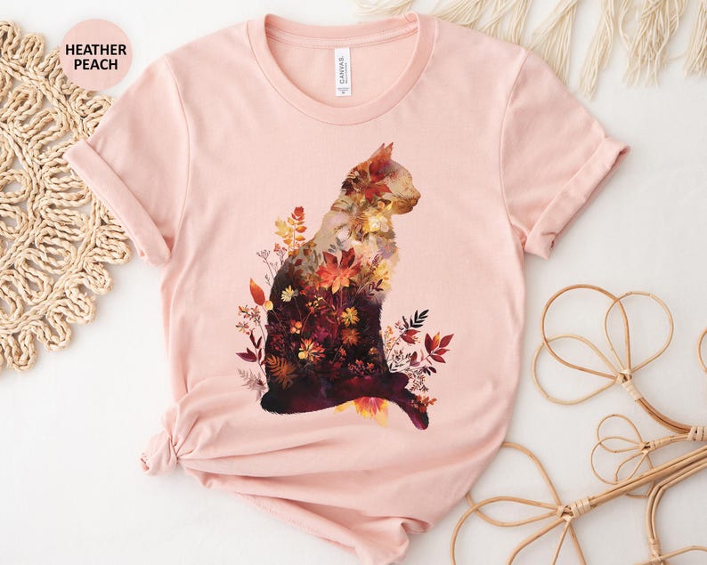 May include: Heather peach t-shirt featuring a cat silhouette filled with autumn flowers and foliage in shades of red, orange, and yellow. The shirt has short sleeves and a crew neck. The text "HEATHER PEACH" is in the upper left corner.