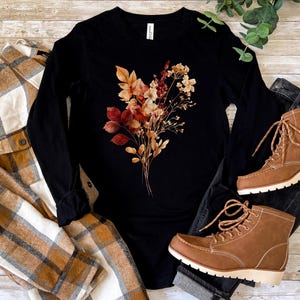 Pu&ograve; includere: Una maglia nera a maniche lunghe con una grafica centrale raffigurante un bouquet di fiori secchi nei toni dell'arancione, del rosso e del marrone. La maglia &egrave; presentata insieme a una camicia a quadri beige e bianca, jeans neri e stivaletti.