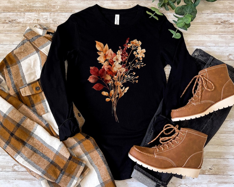 Pu&ograve; includere: Una maglia nera a maniche lunghe con una grafica centrale raffigurante un bouquet di fiori secchi nei toni dell'arancione, del rosso e del marrone. La maglia &egrave; presentata insieme a una camicia a quadri beige e bianca, jeans neri e stivaletti.