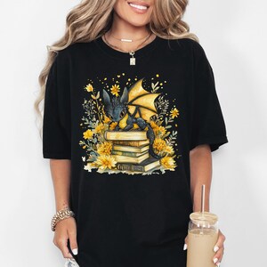 Puede incluir: Camiseta negra con una ilustraci&oacute;n en acuarela de un drag&oacute;n sobre una pila de libros, rodeado de flores y hojas amarillas. El drag&oacute;n tiene escamas negras y alas amarillas. El dise&ntilde;o est&aacute; centrado en la parte delantera.