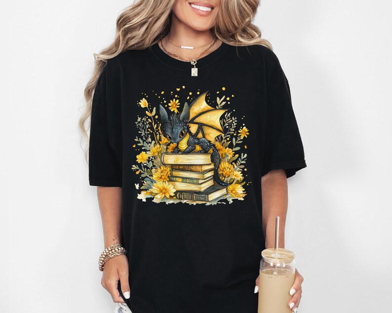 Puede incluir: Camiseta negra con una ilustraci&oacute;n en acuarela de un drag&oacute;n sobre una pila de libros, rodeado de flores y hojas amarillas. El drag&oacute;n tiene escamas negras y alas amarillas. El dise&ntilde;o est&aacute; centrado en la parte delantera.