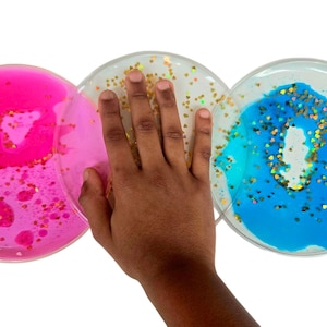 Op de afbeelding: Drie ronde, transparante sensorische speelgoedjes met kleurrijke vloeistof en glitter erin. De speelgoedjes zijn roze, goud en blauw.