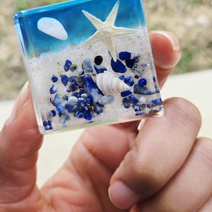 May include: A clear, square resin paperweight featuring a miniature beach scene. The scene includes a white starfish, white sand, blue and gray stones, a seashell, and blue resin representing the ocean and sky.