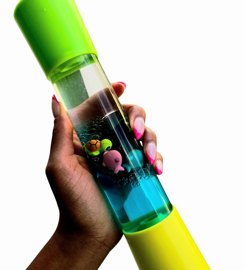 May include: A clear, cylindrical sensory toy with a green and yellow cap and base. The tube contains blue liquid and small, colorful sea creature figurines. The toy is held at an angle against a white background.