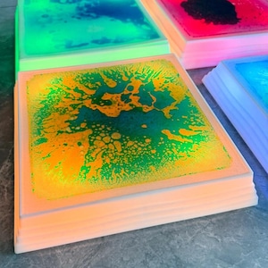 Może przedstawiać: Kwadratowa, biała, podświetlana płytka z zielonym i ż&oacute;łtym płynem w środku. Płyn wiruje i ma bąbelki. Płytka jest podświetlona od dołu, co nadaje jej ciepły blask.