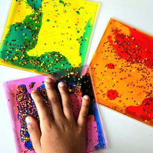 Könnte beinhalten: Drei farbenfrohe sensorische Flüssigkeitsfliesen. Eine Fliese ist gelb und grün, eine andere orange und rot, und die dritte rosa und blau. Jede Fliese enthält Glitzer und wird von einer Hand berührt.