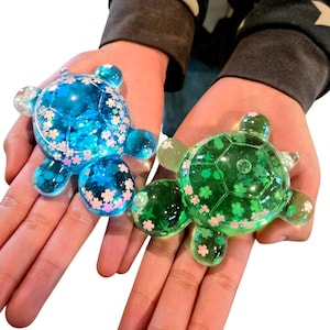 Peut inclure: Deux figurines décoratives en forme de tortue. L'une est bleue avec des motifs floraux blancs et roses, l'autre est verte avec des détails floraux similaires. Les deux sont transparentes et semblent être en résine.