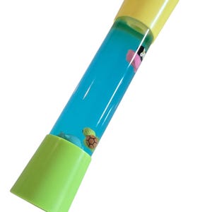 May include: A cylindrical toy with a yellow top and a green base. The clear tube contains blue liquid and small floating figures of a turtle, a dolphin, and a pink flower. The toy is designed to be tilted to watch the figures float.