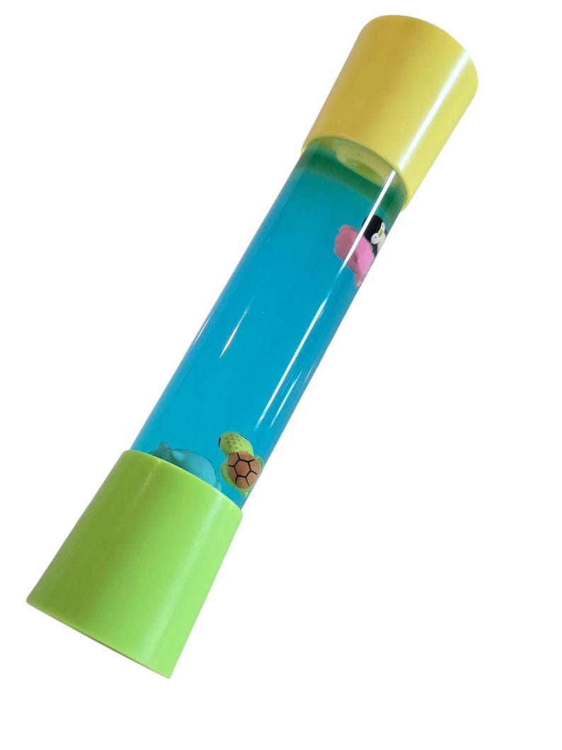 May include: A cylindrical toy with a yellow top and a green base. The clear tube contains blue liquid and small floating figures of a turtle, a dolphin, and a pink flower. The toy is designed to be tilted to watch the figures float.