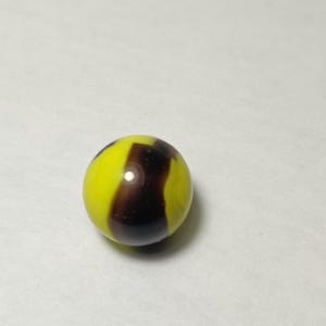 May include: A single, round marble with a yellow base and a dark brown, swirled pattern. The marble is smooth and reflective, with a glossy finish. This classic toy is a collectible item.