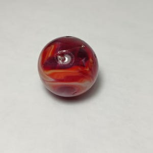 May include: A round, red and orange marbled bead with a hole drilled through the top. The bead has a glossy finish and appears to be made of glass or a similar material. The colors swirl together, creating a unique pattern.