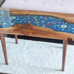 Op de afbeelding: Een rechthoekige houten salontafel met een blauw hars rivierontwerp ingelegd met kleurrijke bloemen. De tafel heeft vier bruine, taps toelopende poten en staat op een wit tapijt. Het hout heeft een natuurlijke nerf.