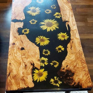 Op de afbeelding: Rechthoekige salontafel met een zwart hars middenstuk waarin gele zonnebloemen zijn ingelegd. De tafel heeft natuurlijke houten randen en staat op vier donkere metalen poten. Het hout heeft een lichtbruine kleur met donkerbruine nerfpatronen.