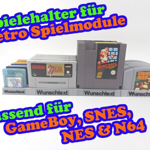 Könnte beinhalten: Ein grauer Plastik-Spielekartuschenhalter mit dem Text "Spielehalter für Retro Spielmodule" und "Wunschtext" darauf. Der Halter ist für Spielekartuschen von GameBoy, SNES, NES und N64 ausgelegt.