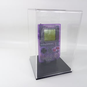 Può includere: Una console per videogiochi portatile Game Boy viola in una teca di acrilico trasparente. La console ha uno schermo in bianco e nero e pulsanti rossi.