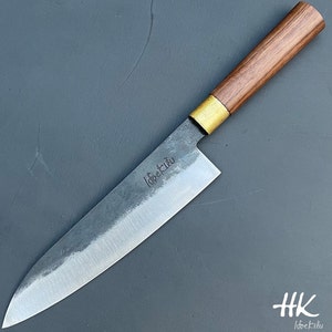 Op de afbeelding: Een lang, scherp, roestvrijstalen keukenmes met een houten handvat en een messing bolster. Het lemmet is gegraveerd met de tekst "Idoekiu".