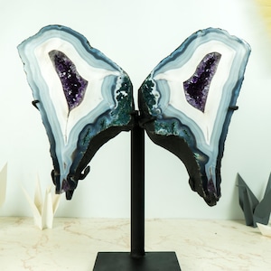Pode incluir: Um par de metades de geoda de ágata com cristais de ametista no interior, montadas em um suporte de metal preto. As metades da geoda são de cor azul claro e branco com bordas verde escuro e preto.