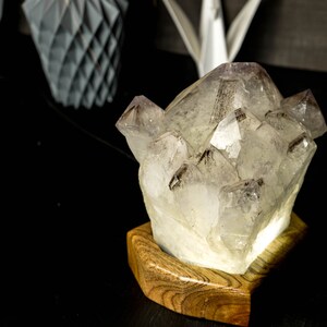 May include: A clear crystal cluster lamp with a wooden base. The crystal is illuminated from within, casting a warm glow. The base is made of dark wood with a natural grain pattern.