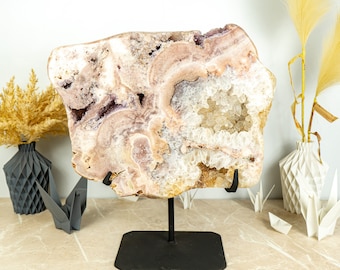 May include: A large, natural amethyst geode with a pink and white colour scheme. The geode is displayed on a black metal stand.