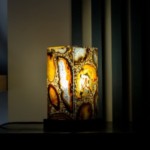 Pode incluir: Um abajur de mesa feito de pedra de ágata natural com um esquema de cores quente, amarelo e marrom. A lâmpada está acesa e projeta um brilho suave.