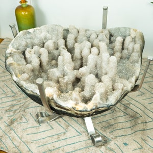 Pode incluir: Uma grande geoda de cristal branco com um exterior áspero cinza escuro. A geoda está montada em um suporte de metal com quatro pernas.