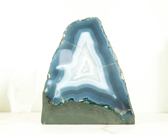 May include: A polished, triangular agate stone with bands of blue, white, and gray. The stone has a dark gray base and a rough, natural edge. This decorative piece is ideal for home decor.