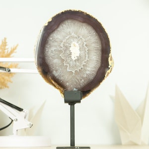 Puede incluir: Una gran rebanada de ágata redonda y blanca con bordes marrones y dorados, expuesta en un soporte de metal negro. La ágata tiene un patrón natural y orgánico.