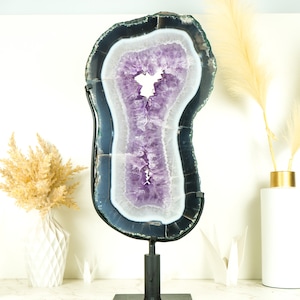 Pode incluir: Uma grande fatia de geoda de ametista com um suporte de metal preto. A geoda é roxa e branca com uma forma natural irregular.