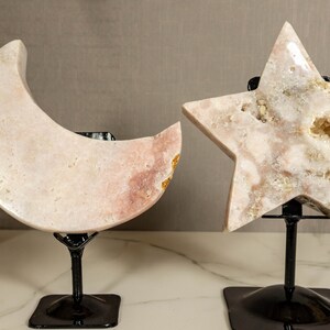 May include: A pink crystal moon and star on black metal stands. The moon and star are carved from a single piece of crystal and have a natural, organic look. The crystal is translucent and has a slight pink hue. The stands are simple and elegant, and they allow the crystal to be displayed in a variety of ways.