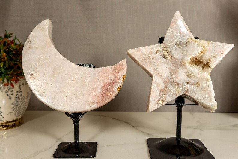 May include: A pink crystal moon and star on black metal stands. The moon and star are carved from a single piece of crystal and have a natural, organic look. The crystal is translucent and has a slight pink hue. The stands are simple and elegant, and they allow the crystal to be displayed in a variety of ways.