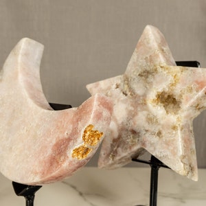 May include: Two pink and white crystal sculptures, one shaped like a crescent moon and the other like a star, are displayed on black metal stands. Both sculptures have gold flecks throughout.