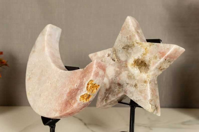 May include: Two pink and white crystal sculptures, one shaped like a crescent moon and the other like a star, are displayed on black metal stands. Both sculptures have gold flecks throughout.