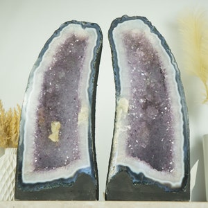 Op de afbeelding: Twee grote amethistgeoden, elk met een paars en wit kristallen interieur, worden getoond op een wit oppervlak. De geoden zijn ongeveer 30 cm hoog en 20 cm breed.