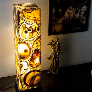 Pode incluir: Candeeiro de mesa retangular com um design único. O candeeiro apresenta uma pedra translúcida de cor âmbar com padrões intrincados. O candeeiro está iluminado, mostrando as variações naturais da pedra. Está sobre uma superfície escura.
