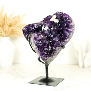 Pode incluir: Uma grande geoda de ametista com uma cor roxa profunda e uma formação de cristal natural. A geoda é exibida em um suporte de metal preto.
