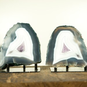 May include: Two agate bookends with a white and gray banded pattern and a purple amethyst crystal in the center. The bookends are on a wooden surface and each has a metal stand.