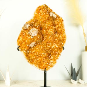 May include: A large, orange citrine crystal cluster on a black metal stand. The crystal is a natural, raw formation with a rough, textured surface.