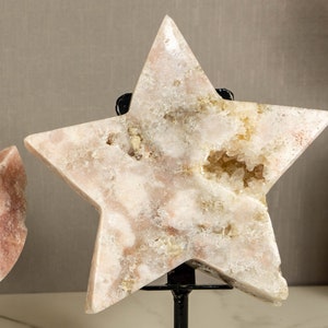 May include: A large, white, star-shaped crystal with a pink hue and a natural, rough texture. The crystal is on a black metal stand.