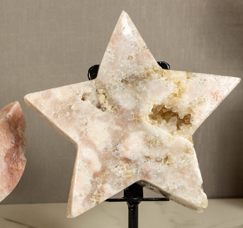 May include: A large, white, star-shaped crystal with a pink hue and a natural, rough texture. The crystal is on a black metal stand.