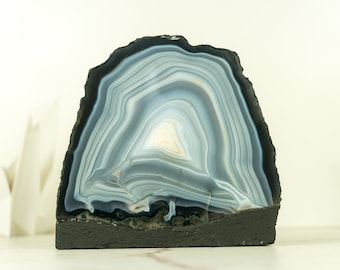 May include: A large, gray and black agate geode with a natural, rough edge. The geode is sliced in half, revealing the intricate, banded pattern of the agate. The geode is mounted on a black base.