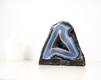 May include: A blue and black geode crystal with a triangular shape. The geode has layers of blue and white bands surrounding a dark, crystal-filled center. The exterior is a rough, dark gray.