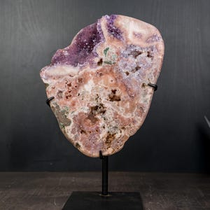 May include: A large, irregularly shaped amethyst geode specimen, mounted on a black metal stand. The geode features a mix of purple, pink, and white hues with visible crystal formations and cavities. The overall aesthetic is natural and decorative.